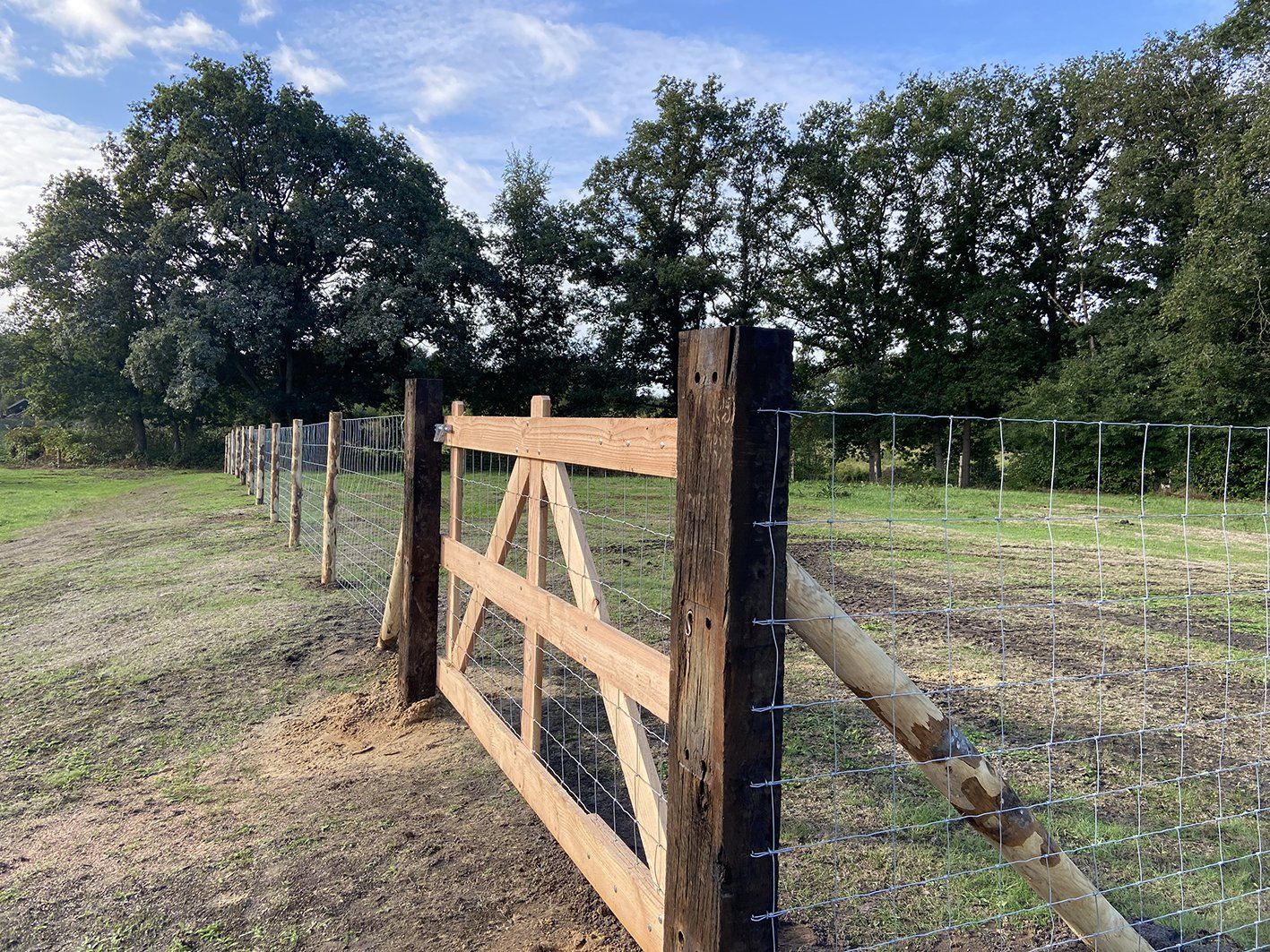 Houten poort en hek in een grasveld, bomen op de achtergrond, onder een blauwe hemel.