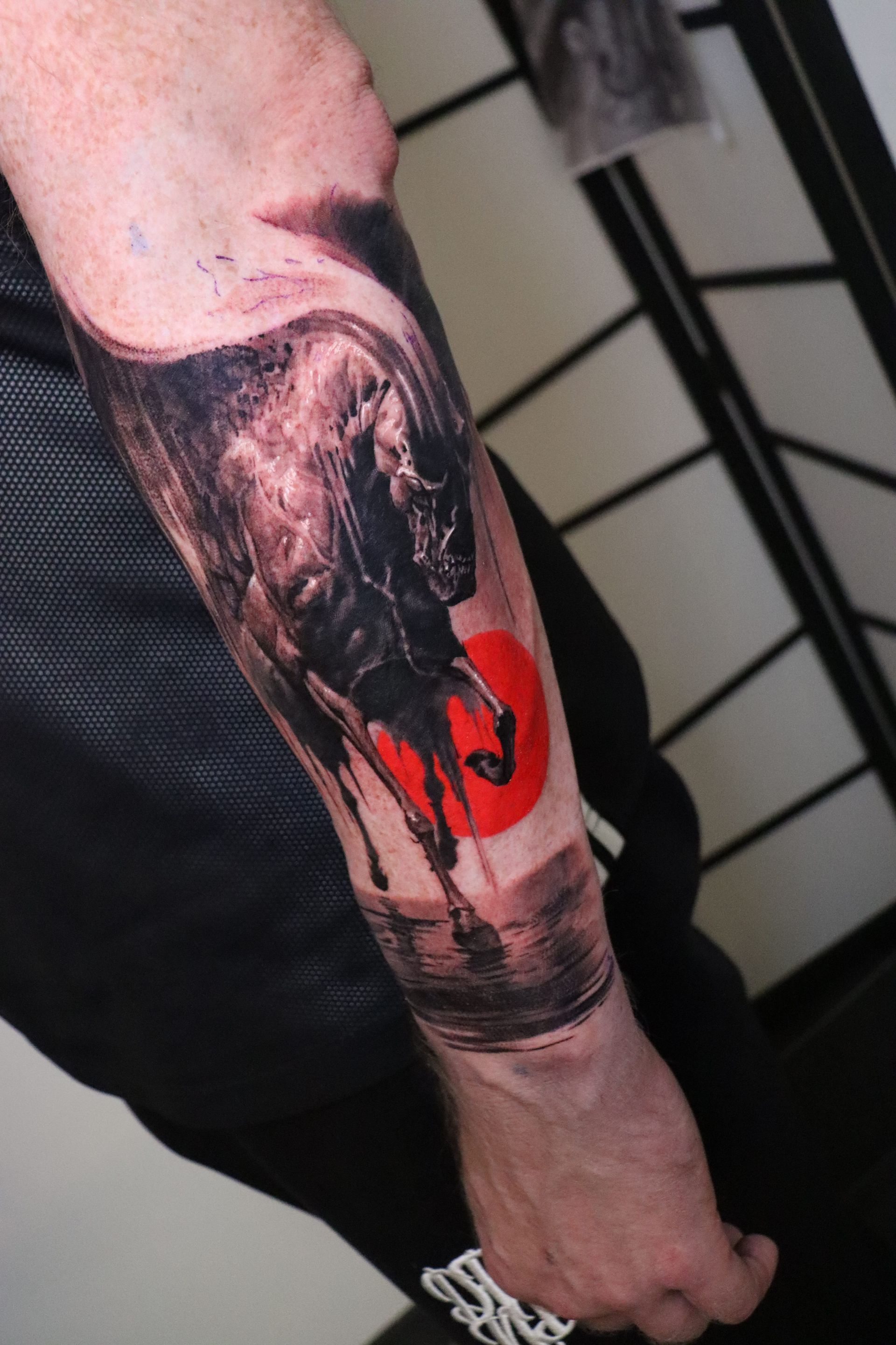 A forearm tattoo of a black bull silhouette against a vibrant red sun, featuring dark, drip-like ink shading.