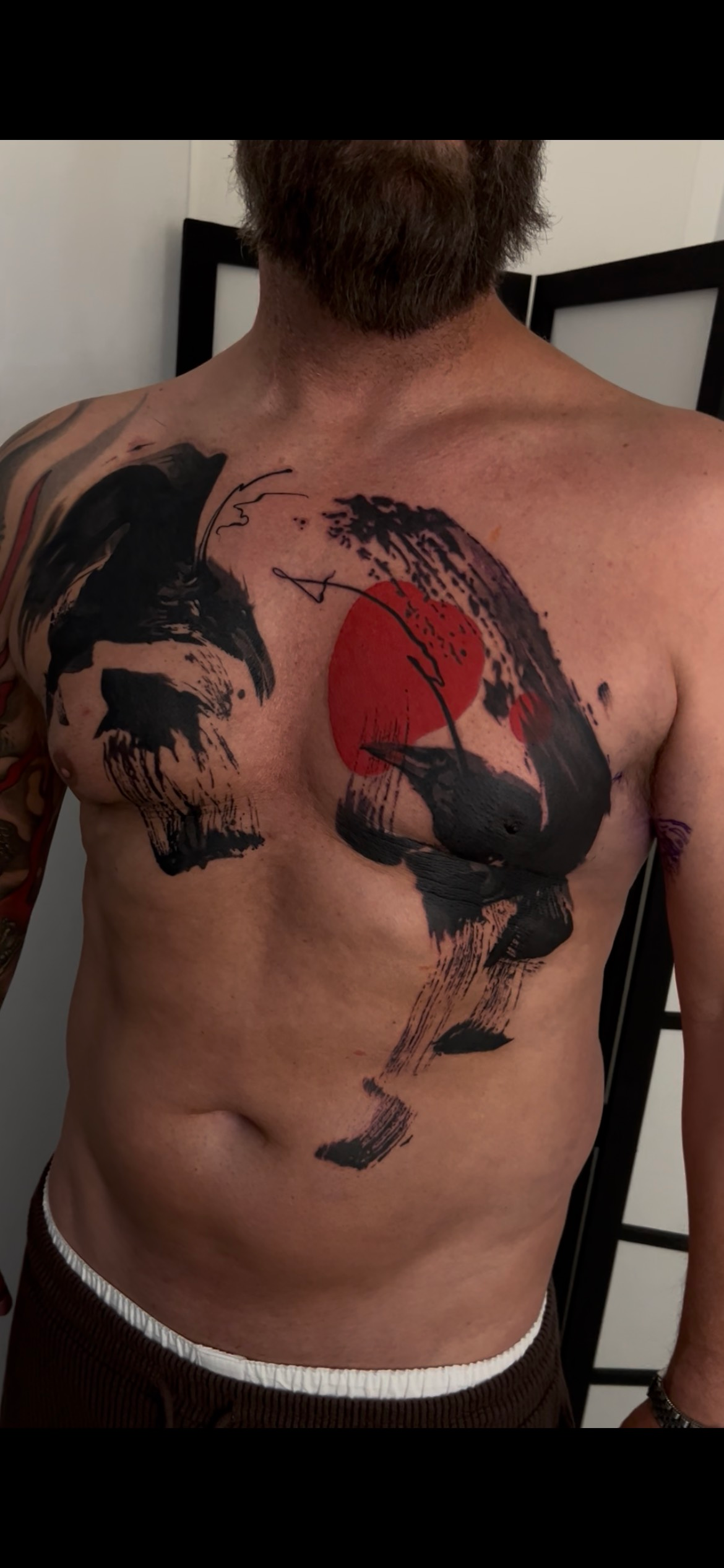 A person’s upper torso featuring a large, abstract black ink-brush style tattoo with a central, vibrant red sun circle.