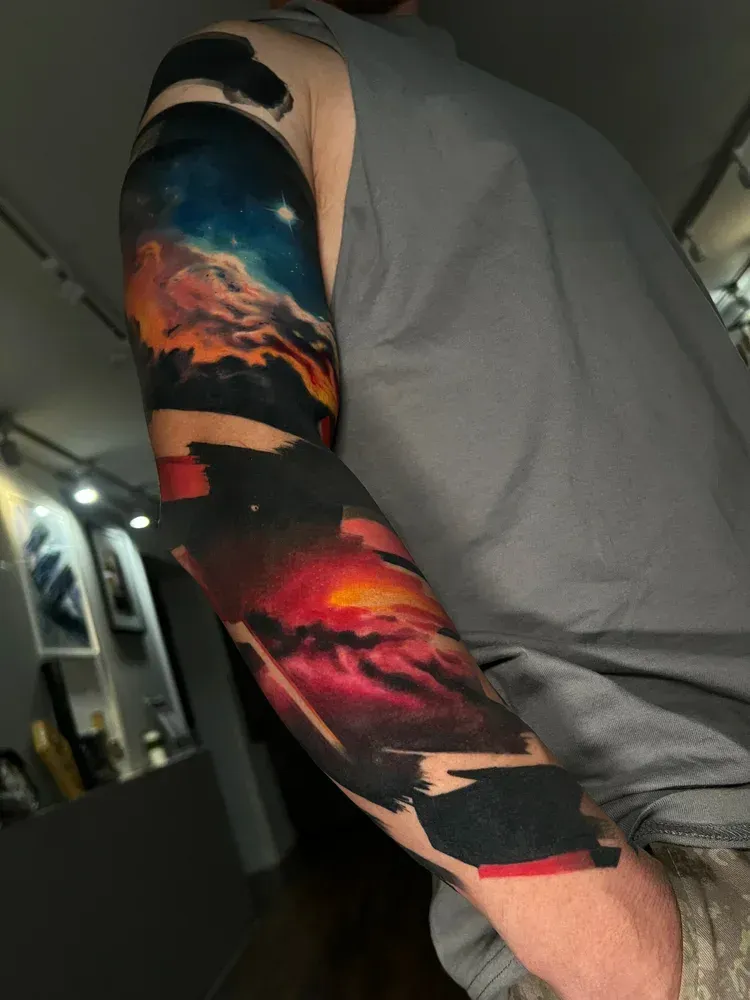 A person’s arm featuring a vibrant, abstract sleeve tattoo with colorful nebulae and bold black ink brushstrokes.