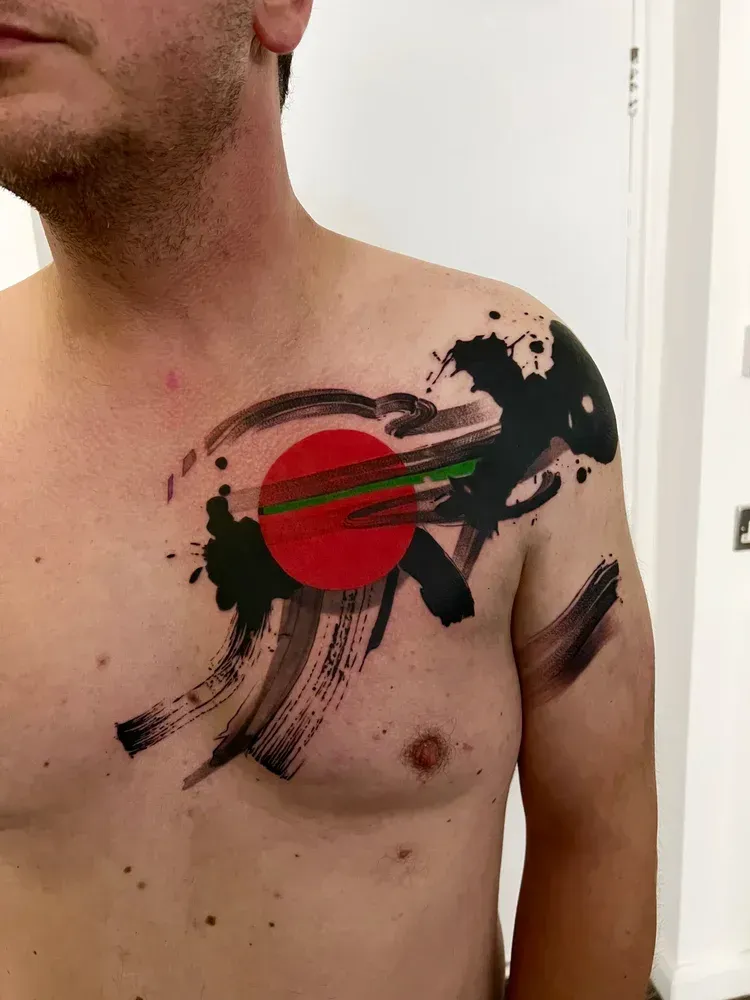 A tattoo on a person's chest and shoulder featuring a large red circle surrounded by black abstract brushstrokes.