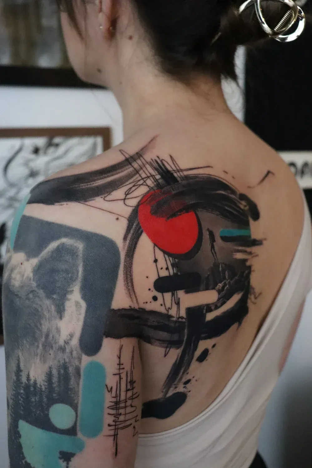 Abstract tattoo on a shoulder featuring black brushstrokes, a solid red circle, and a landscape scene with turquoise shapes.
