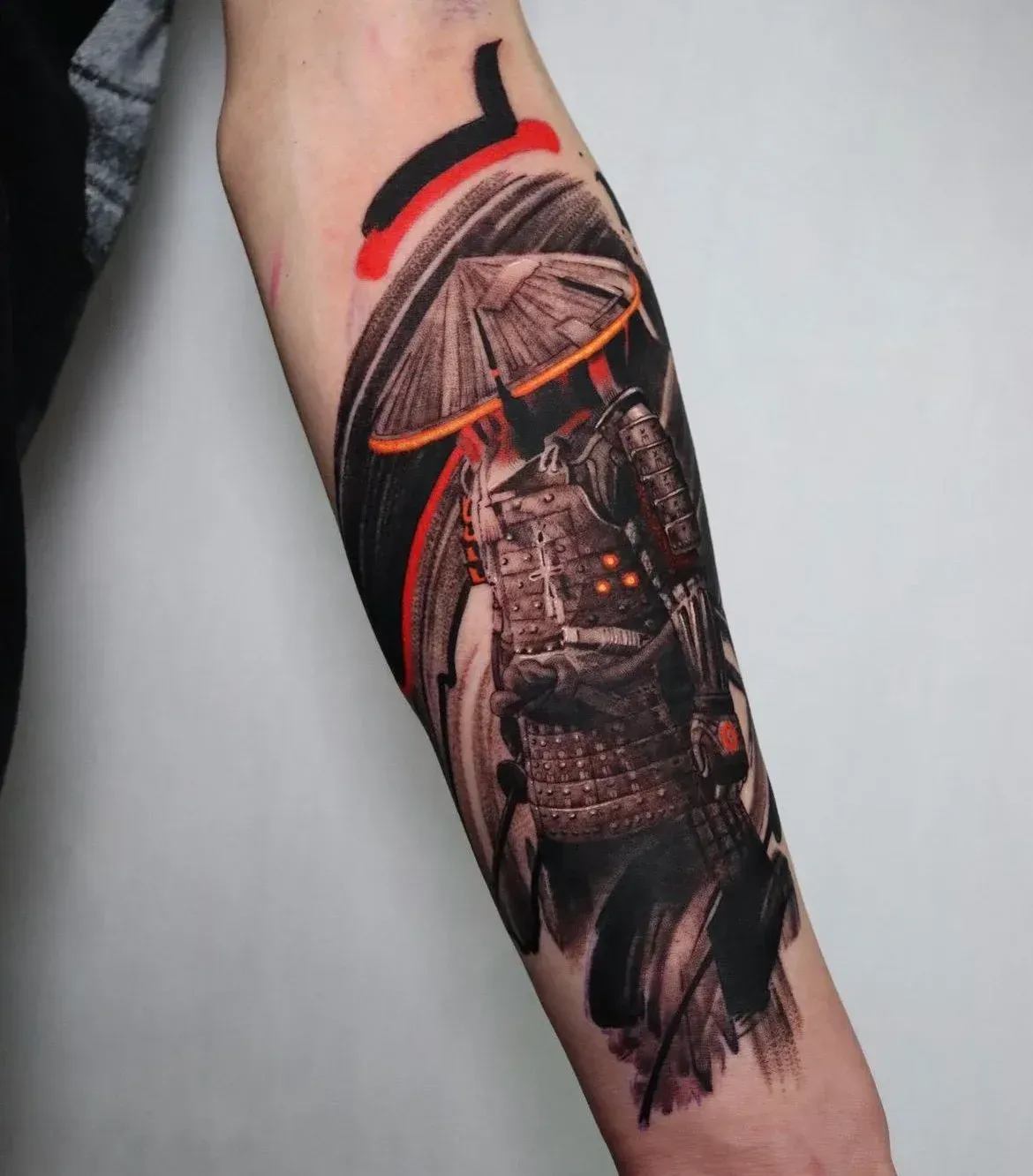 A forearm tattoo of a samurai wearing traditional armor and a wide conical hat, styled with bold black and red ink strokes.