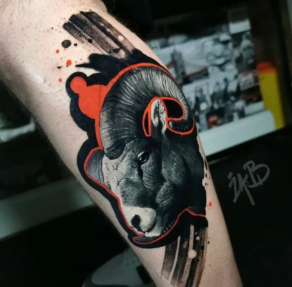 A black-and-grey tattoo of a ram's head on a person's arm, featuring stylized red outlines and brushstroke effects.