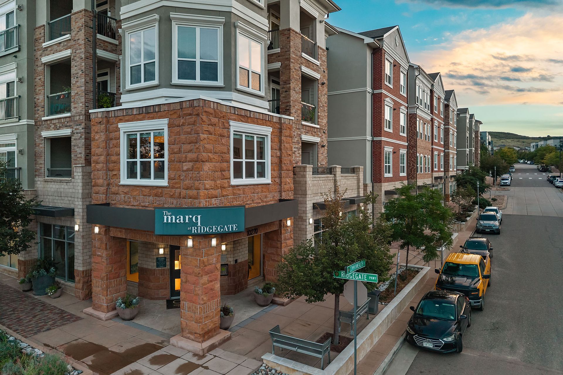Brick apartment building on a city street at dusk, with parked cars and a corner entrance at The Marq at Ridgegate in Lone Tree, CO.