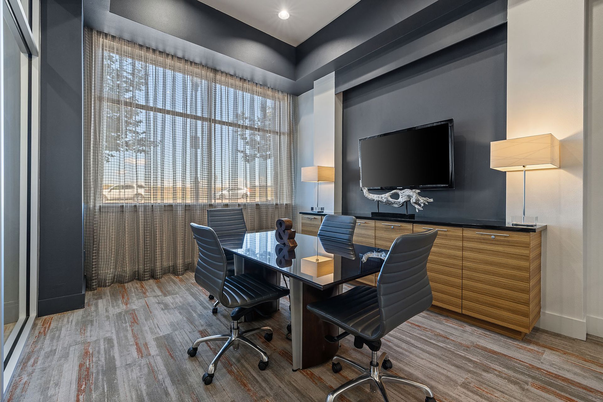 Modern conference room with glass table, black chairs, wall-mounted TV, and large window with curtains at The Marq at Ridgegate in Lone Tree, CO.