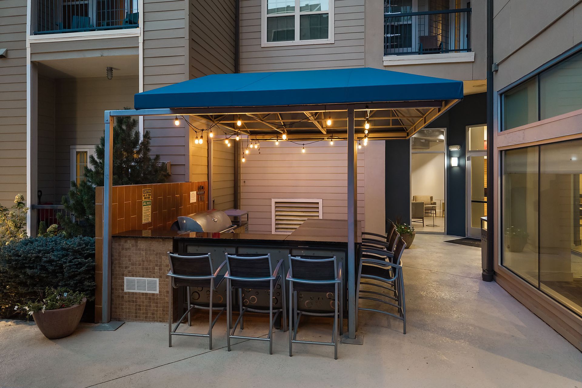 Covered outdoor patio seating area with blue awning, string lights, and barstools beside an apartment building at The Marq at Ridgegate in Lone Tree, CO.