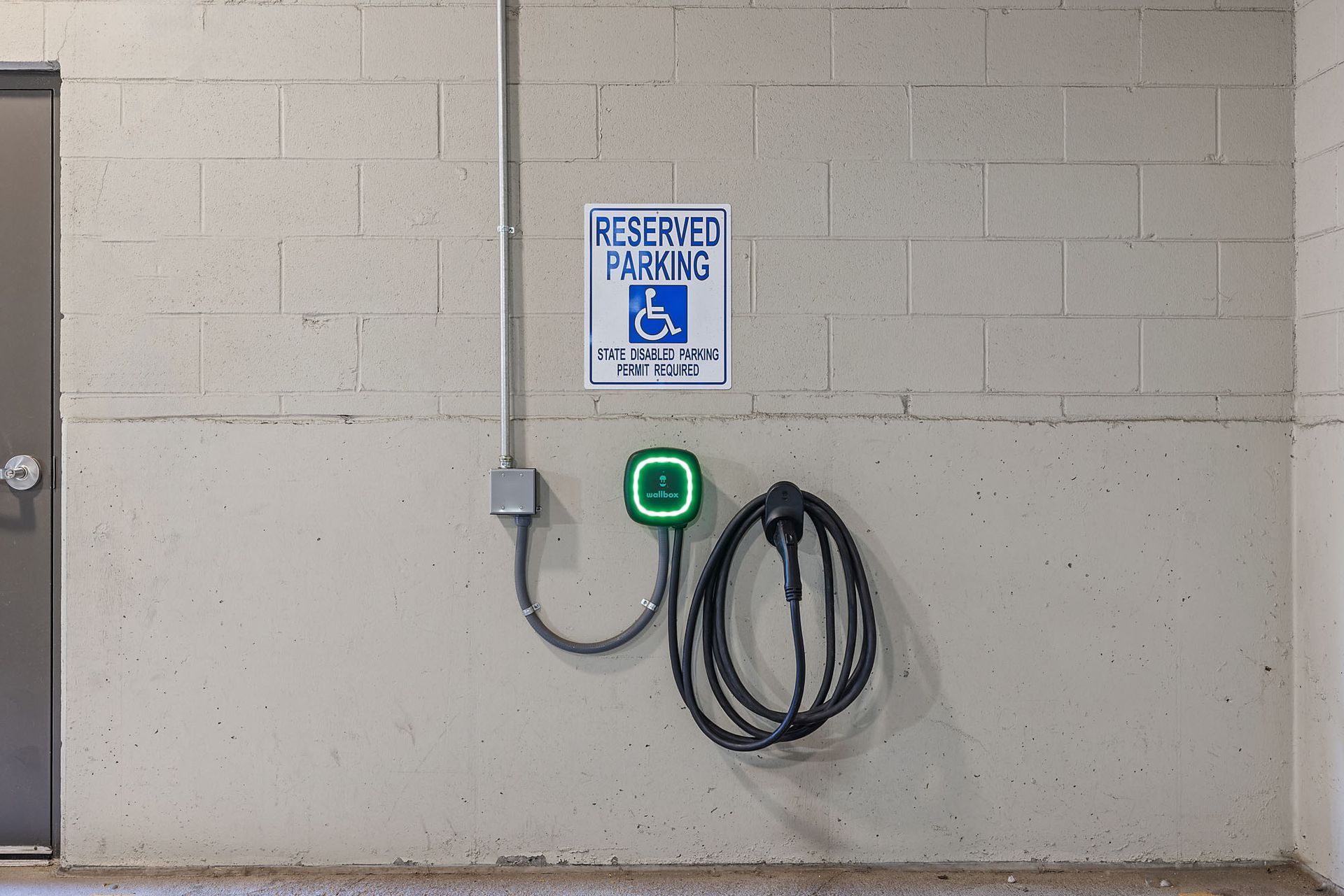 Accessible parking sign on beige wall with electrical outlet, green charger, and coiled black cable nearby at The Marq at Ridgegate in Lone Tree, CO.