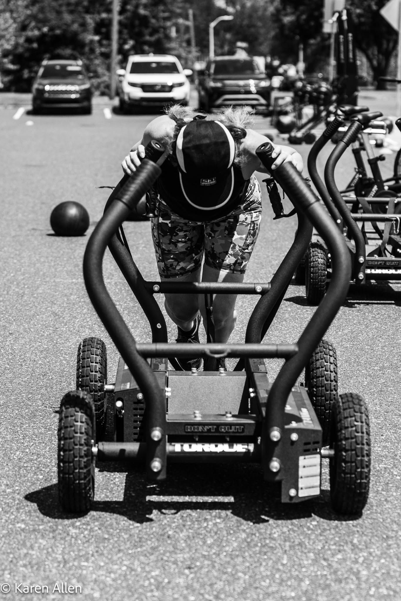 Person riding a low, wheeled cart on a street, with parked cars in the background