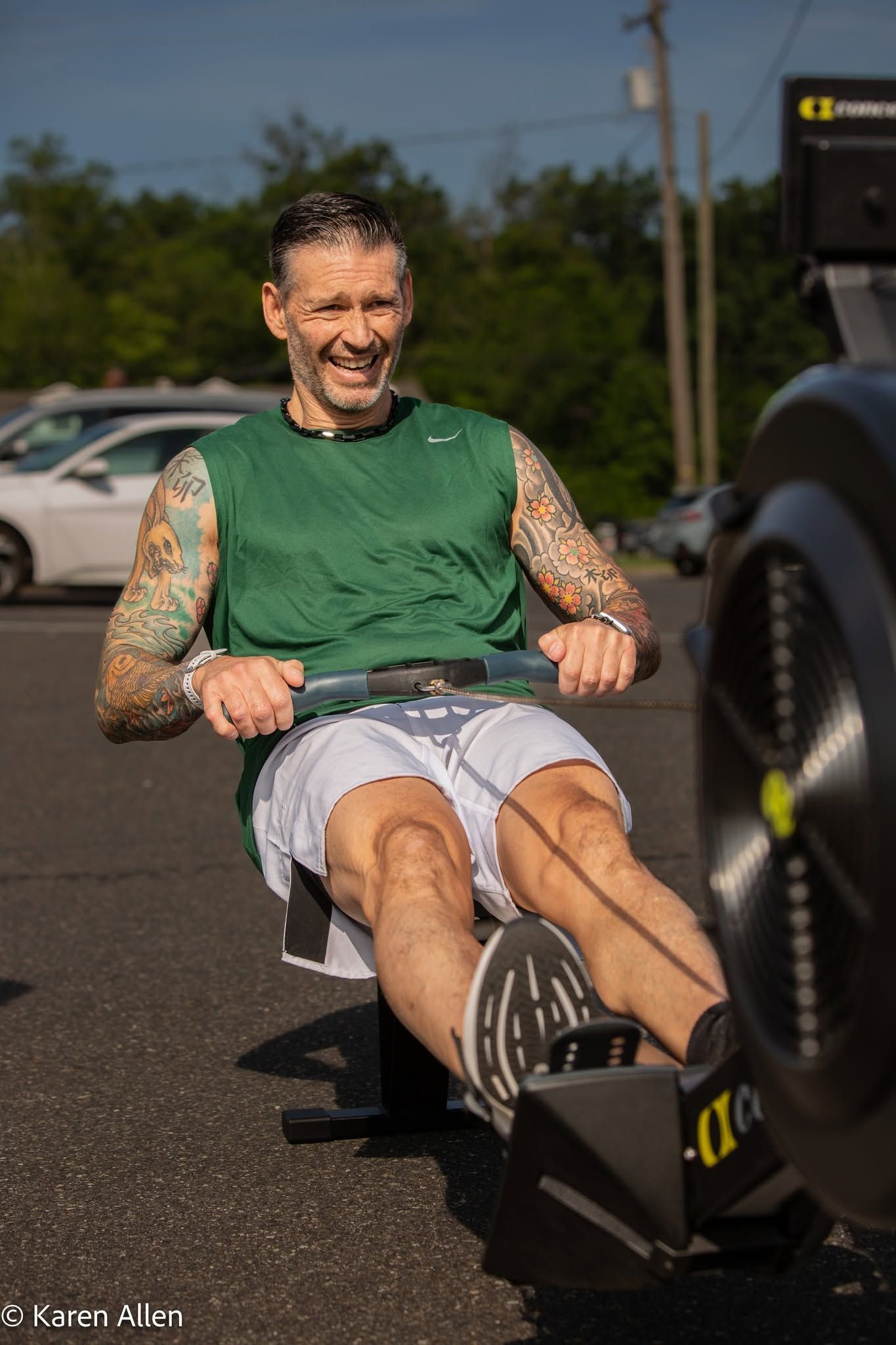 Man rowing on an indoor rowing machine outdoors, wearing a green shirt and gray shorts.