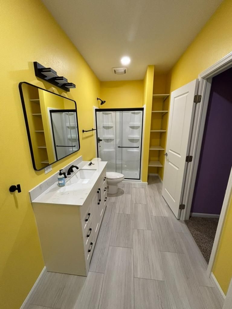 Bright yellow bathroom with white vanity, large mirror, shower, and open doorway with purple room beyond