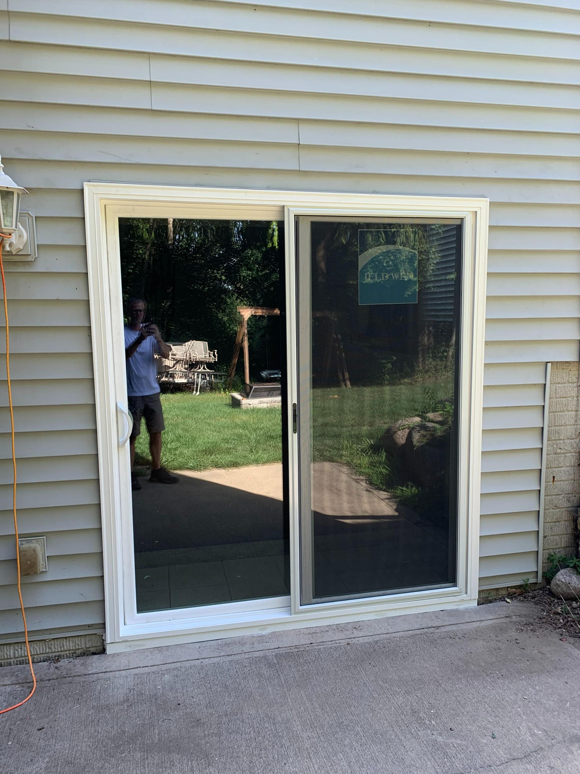 Sliding glass door in beige siding reflecting a backyard and a person standing nearby.