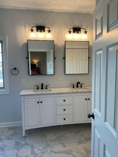 White bathroom vanity with double sinks, mirrors, lights, and a tiled floor, seen through a partially open door
