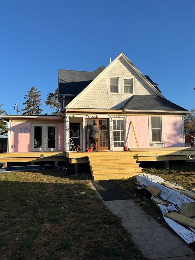 Front view of a pink house with a yellow porch, front steps, and exposed construction materials under a blue sky