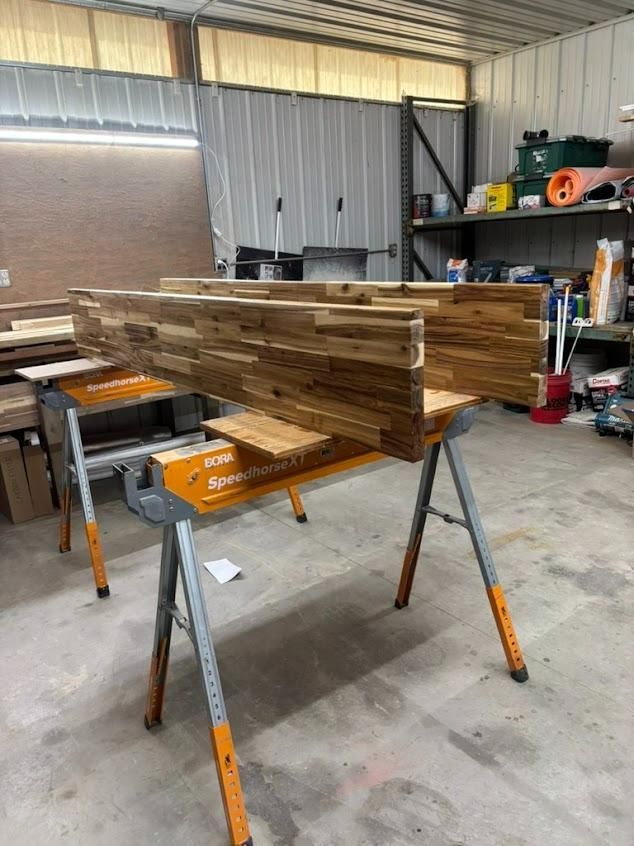 Stacked wooden boards on sawhorses in a workshop with tools and shelves in the background