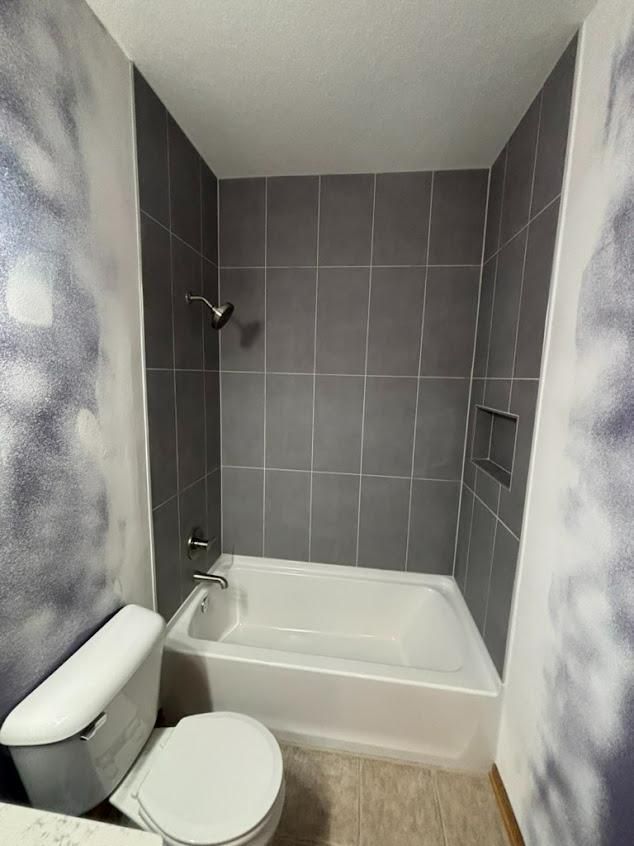 Small bathroom with a white tub, toilet, gray tiled shower, and marbled walls.