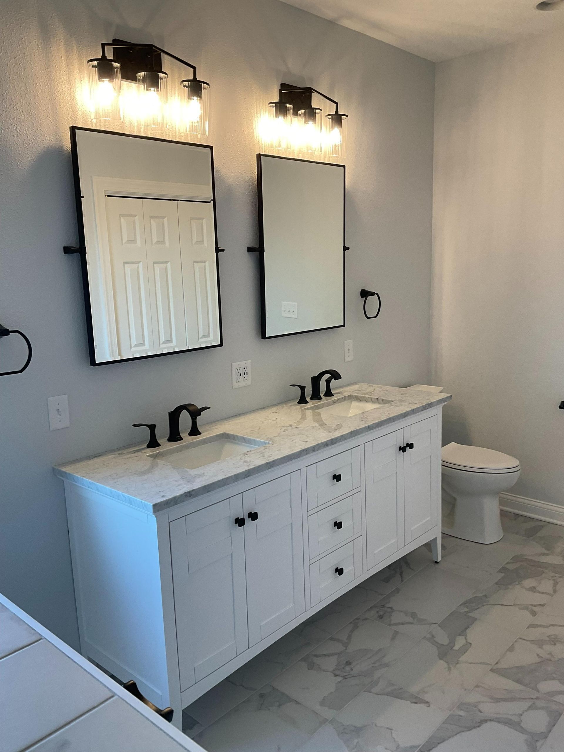 Modern bathroom with double vanity, two mirrors, sconces, and a toilet on marble-pattern floor.