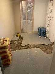 Damaged floor in a small room with exposed dirt, stacked bags, and a blue machine near a plastic-covered door.