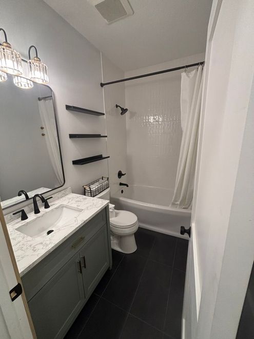 Small bathroom with vanity, toilet, black shelves, and shower with white curtain and dark tile floor.