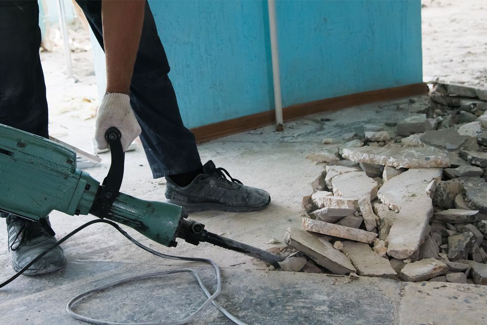 Worker using a jackhammer to break concrete rubble beside a blue wall