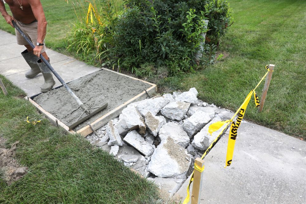 Worker pouring fresh concrete beside a broken sidewalk section marked with caution tape