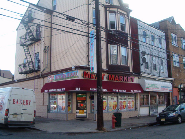Corner bakery and market storefront with delivery truck, brick building, and parked car on a city street