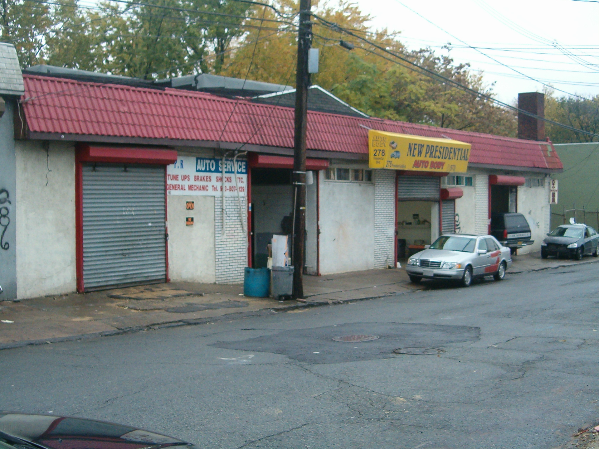 Closed storefronts with metal shutters, red roof, and parked cars along a street