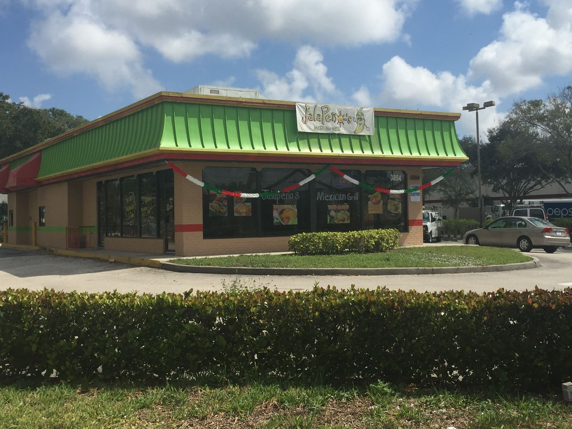 Corner restaurant with green awning and red trim in a parking lot, viewed from across a hedge.