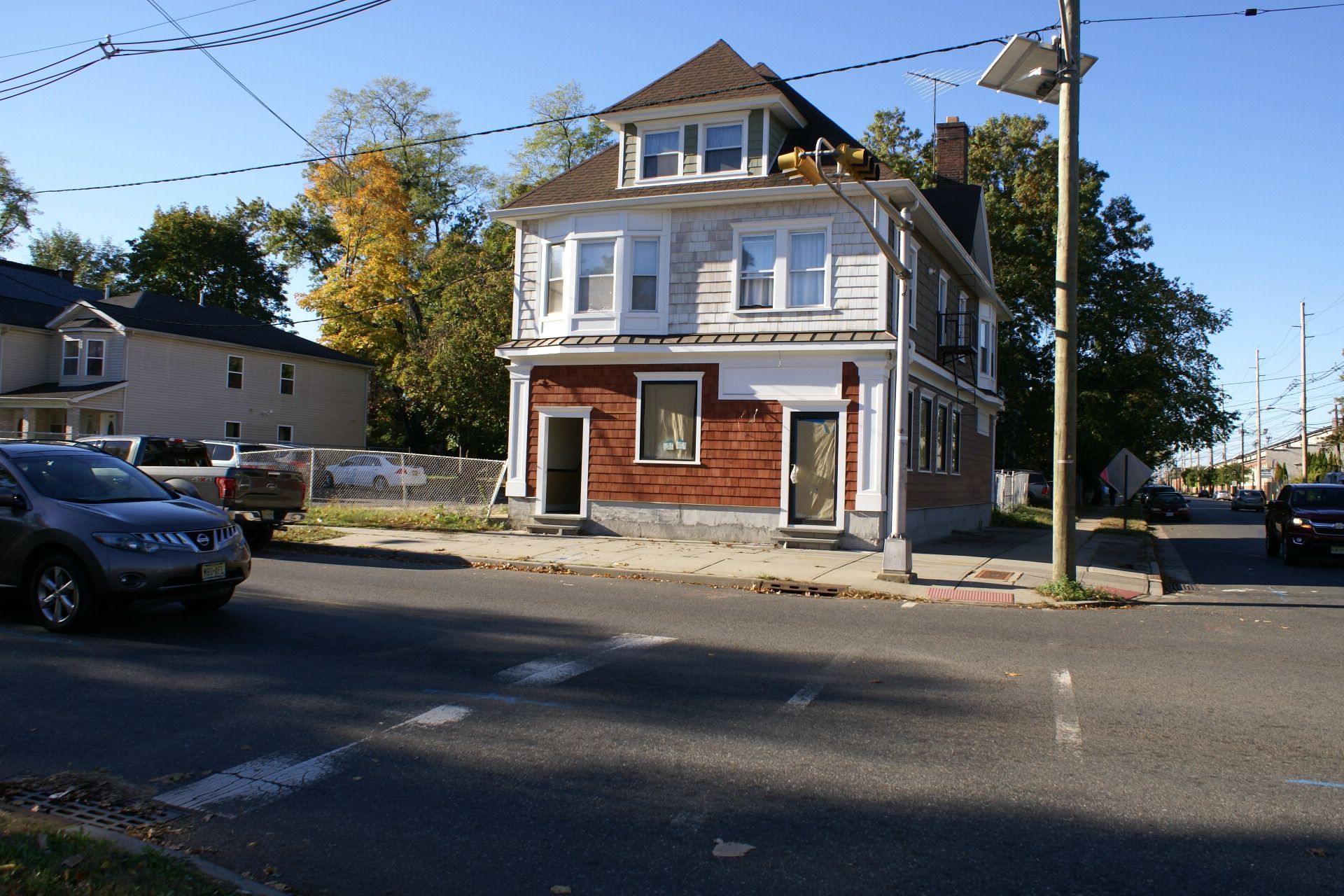 Two-story weathered house on a street corner, with parked cars and utility poles nearby.
