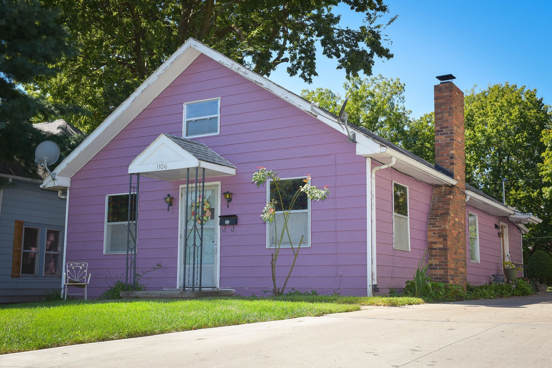 Small purple house with white trim and a brick chimney under trees on a sunny day