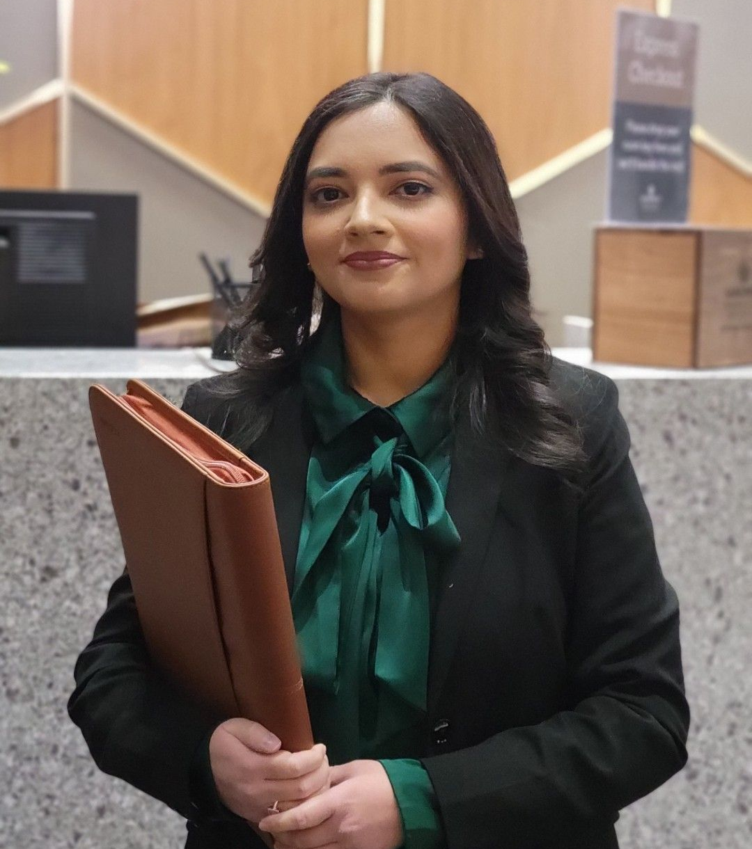 Woman in a black blazer holding a brown folder, standing indoors beside a reception counter.