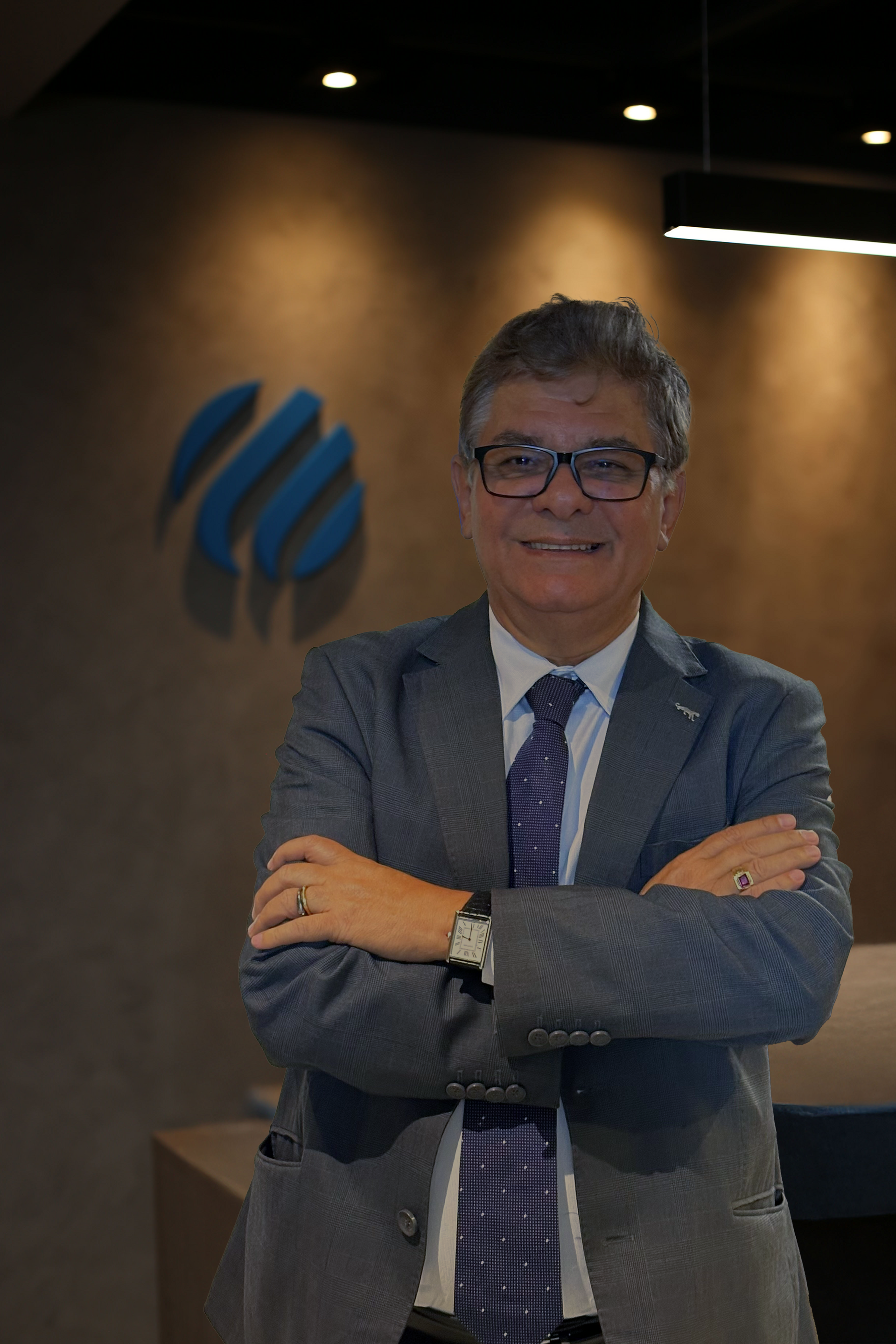 Homem sorridente de terno cinza, com os braços cruzados, em pé no saguão de um escritório com um logotipo azul na parede.