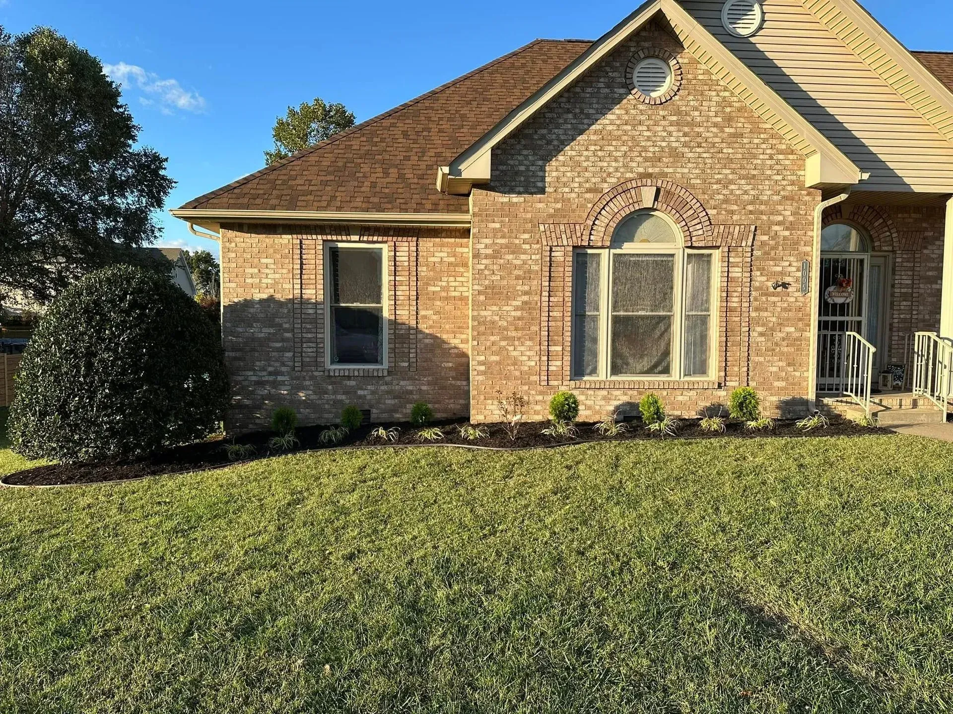 A one-story brick house with a brown shingled roof, arched windows, and a manicured front lawn under a blue sky.