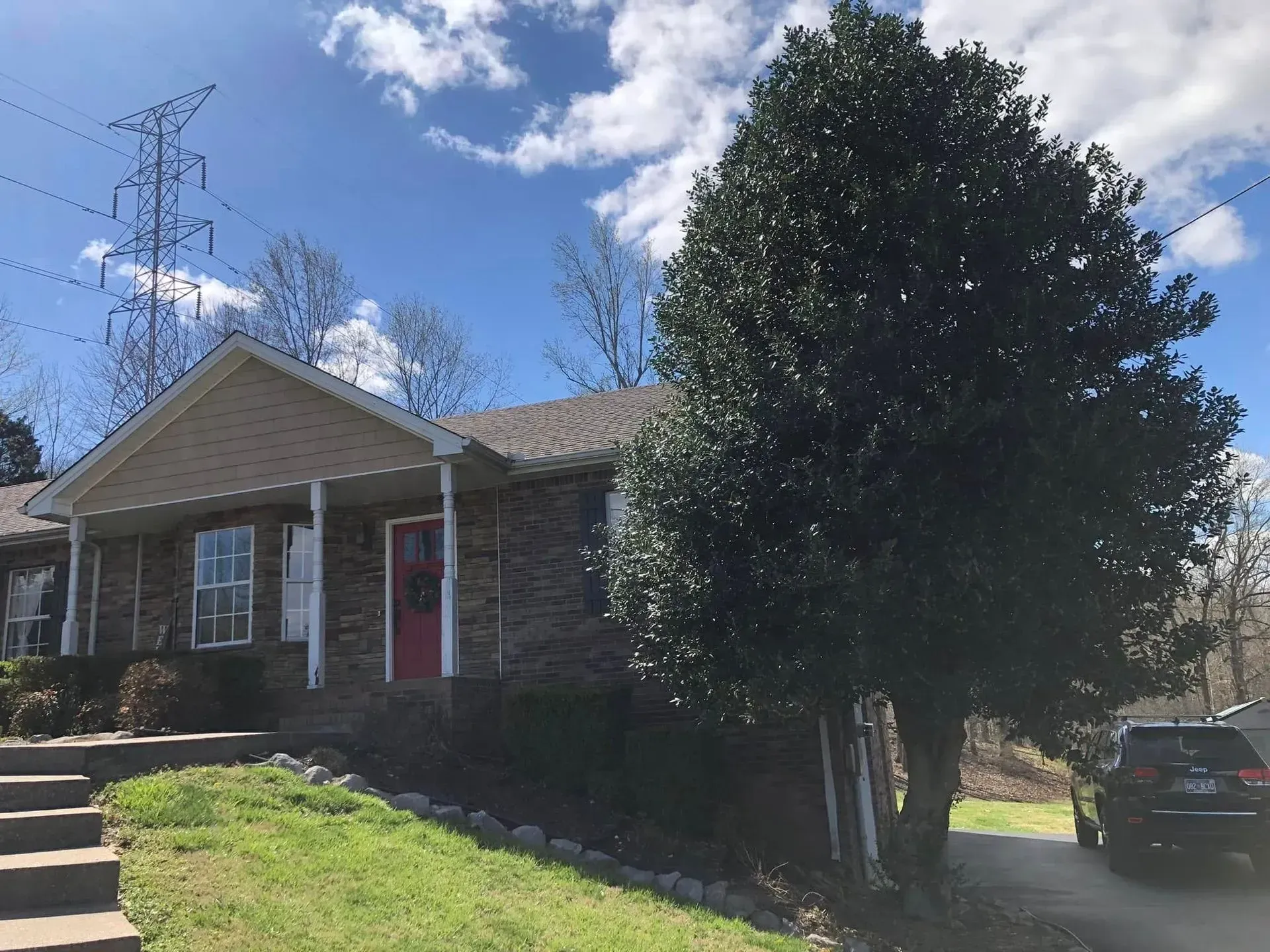 A one-story brick house with a front porch, a red door, and a large evergreen tree in the yard under a blue sky.
