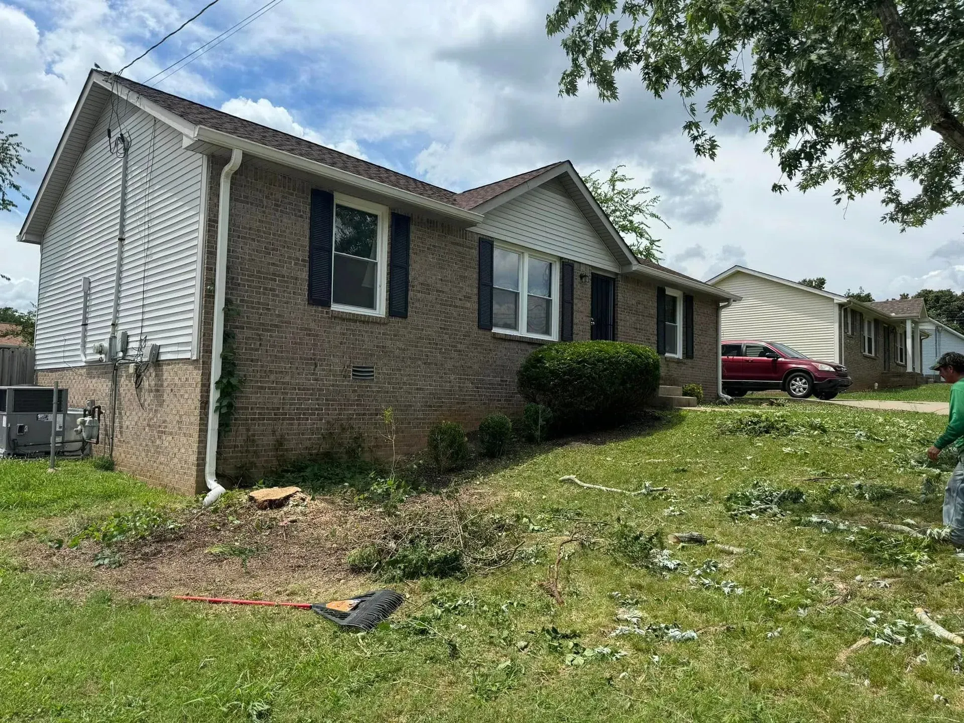 A brick, single-story house with white siding under a cloudy sky, with debris from landscaping scattered on the lawn.