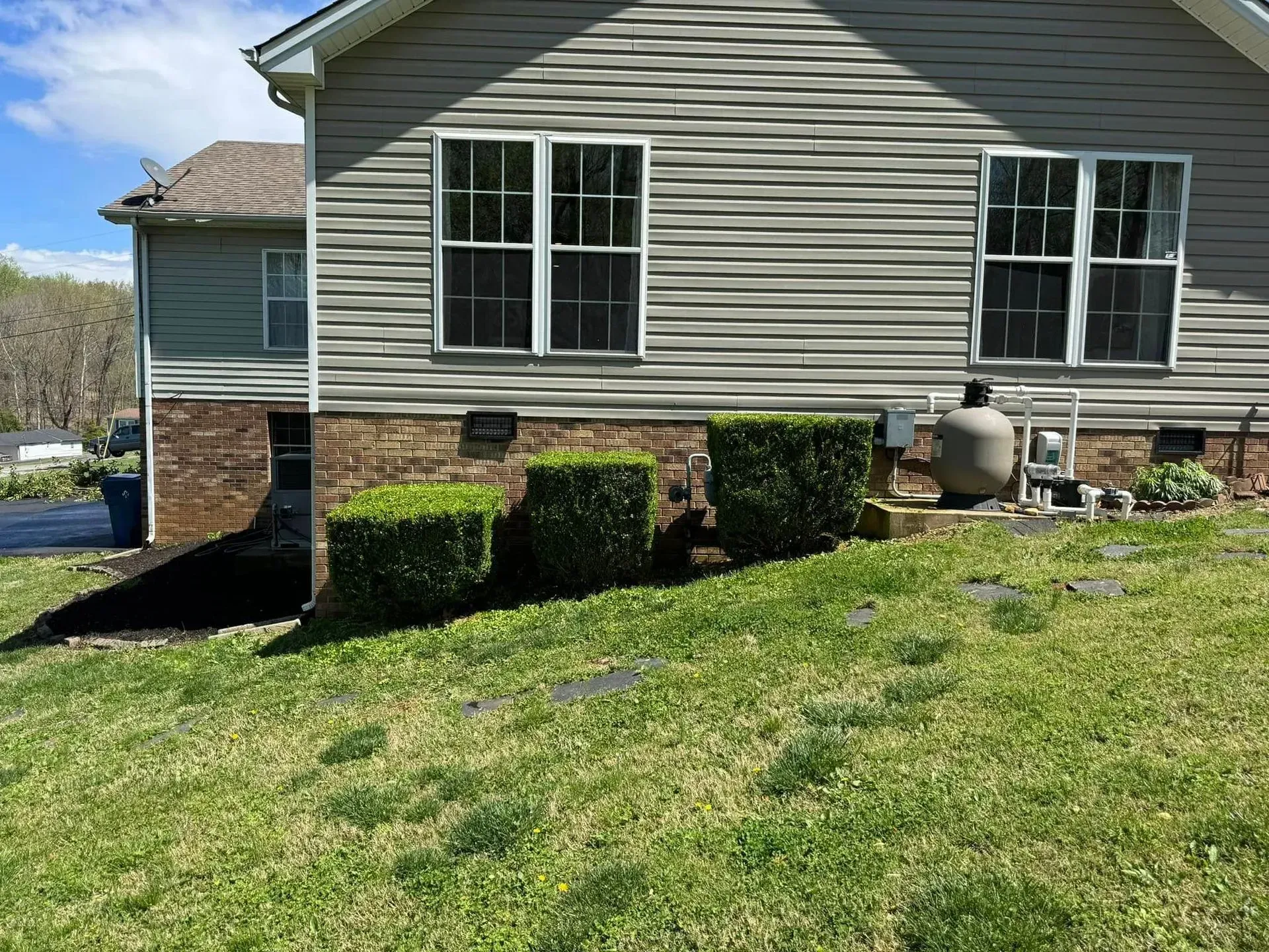 Side view of a beige-sided house with a brick foundation, green bushes, and a propane tank on a grassy lawn.