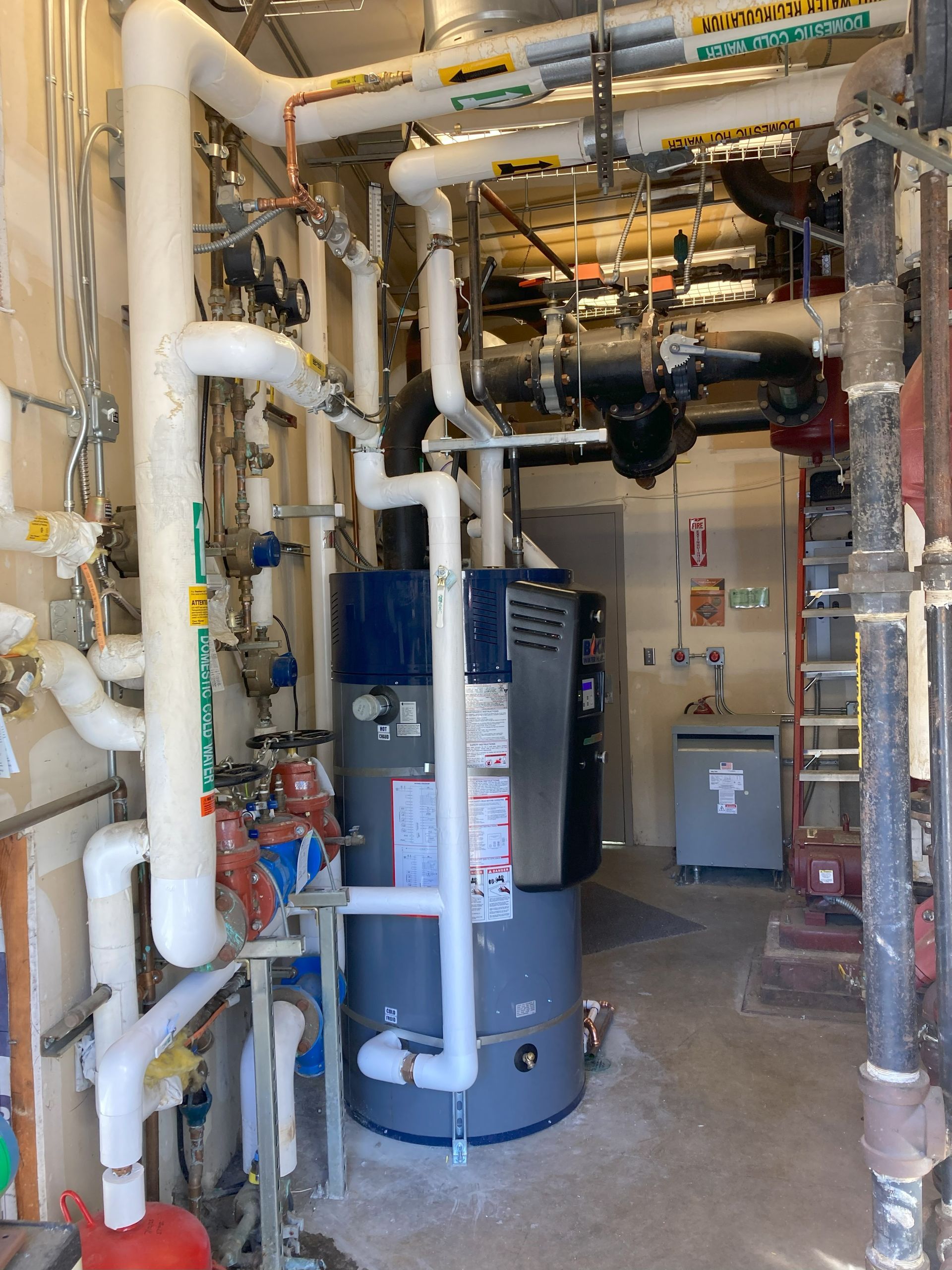 Utility room with large pipes, valves, and a blue water tank in an industrial basement.