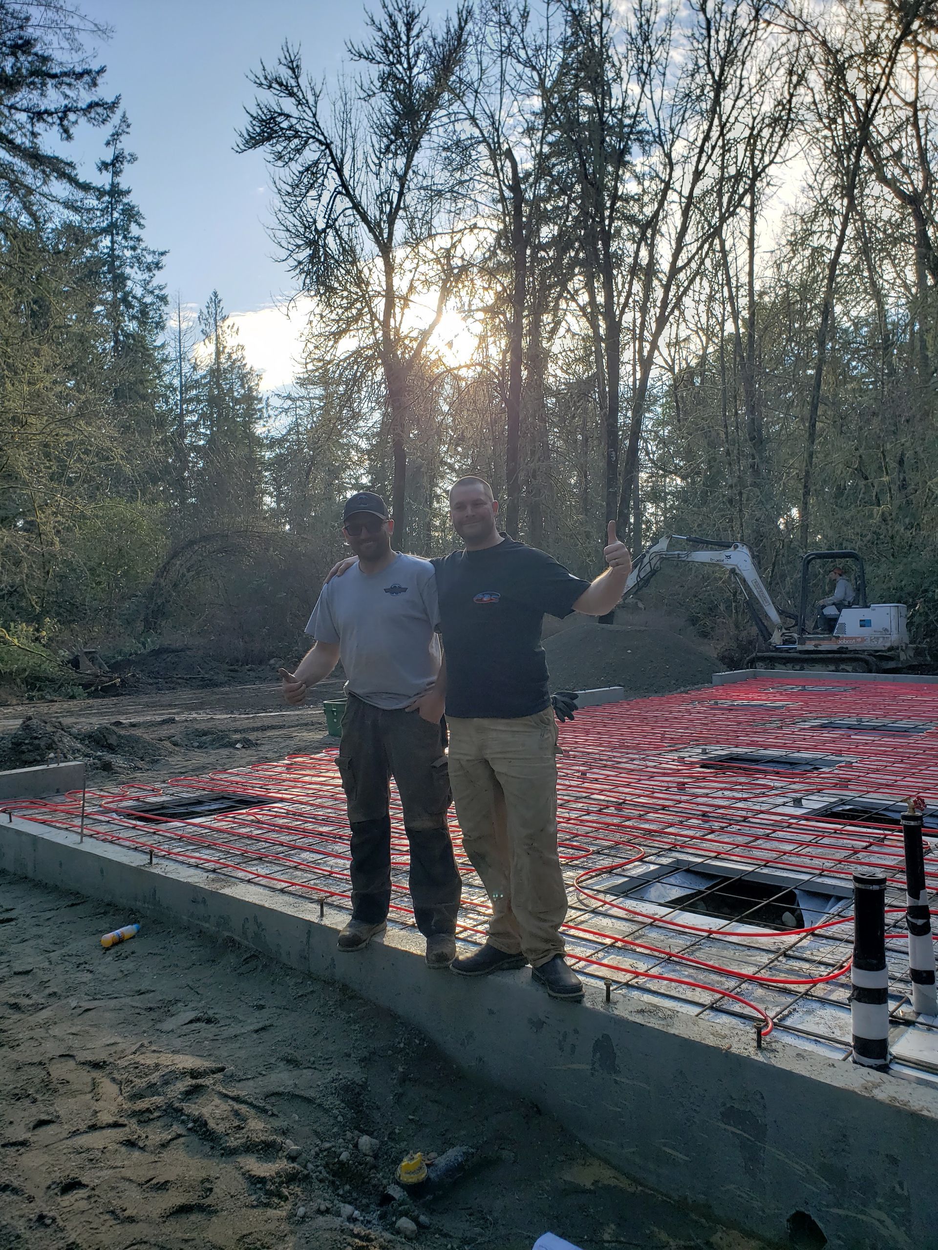 Two people standing on a concrete slab with red pipes and rebar in a wooded construction site at sunset