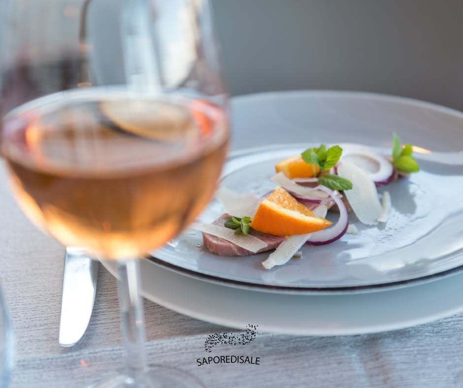 Un bicchiere di vino rosato in primo piano accanto a un piatto di fette d'arancia, formaggio e cipolle rosse.