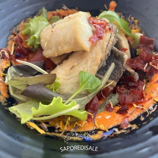 Primo piano di un piatto di pesce impiattato con filetti fritti, salsa, verdure e guarnizione in una ciotola scura.