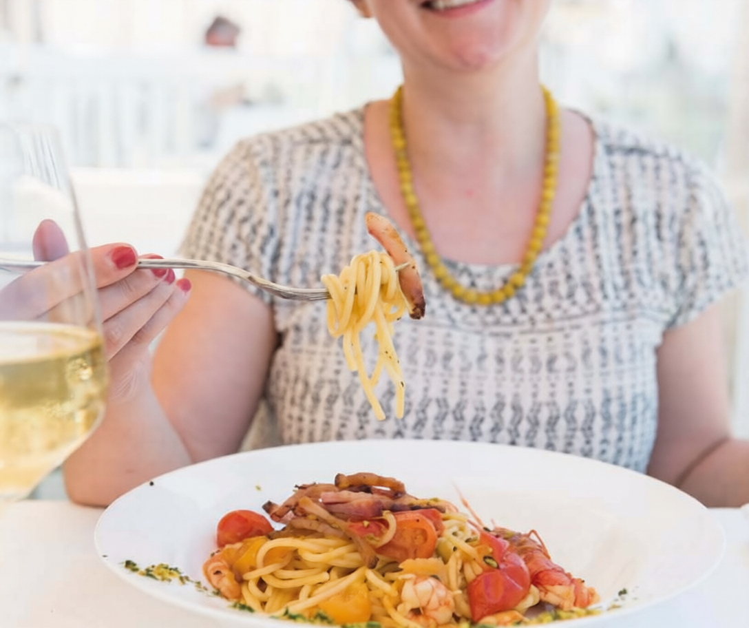 Donna che mangia pasta con pomodori e frutti di mare, sorridente. Tiene in mano una forchetta con la pasta. Vino bianco.