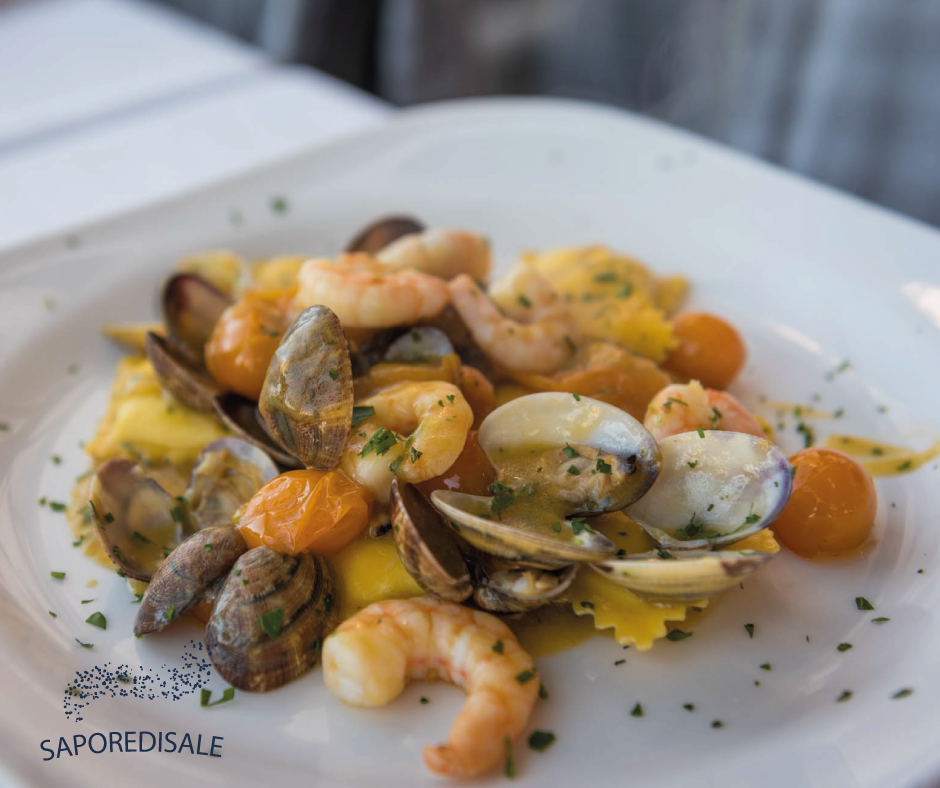 Piatto di pasta ai frutti di mare: gamberi, vongole e ravioli in una salsa leggera, guarnita con prezzemolo e pomodori arancioni.