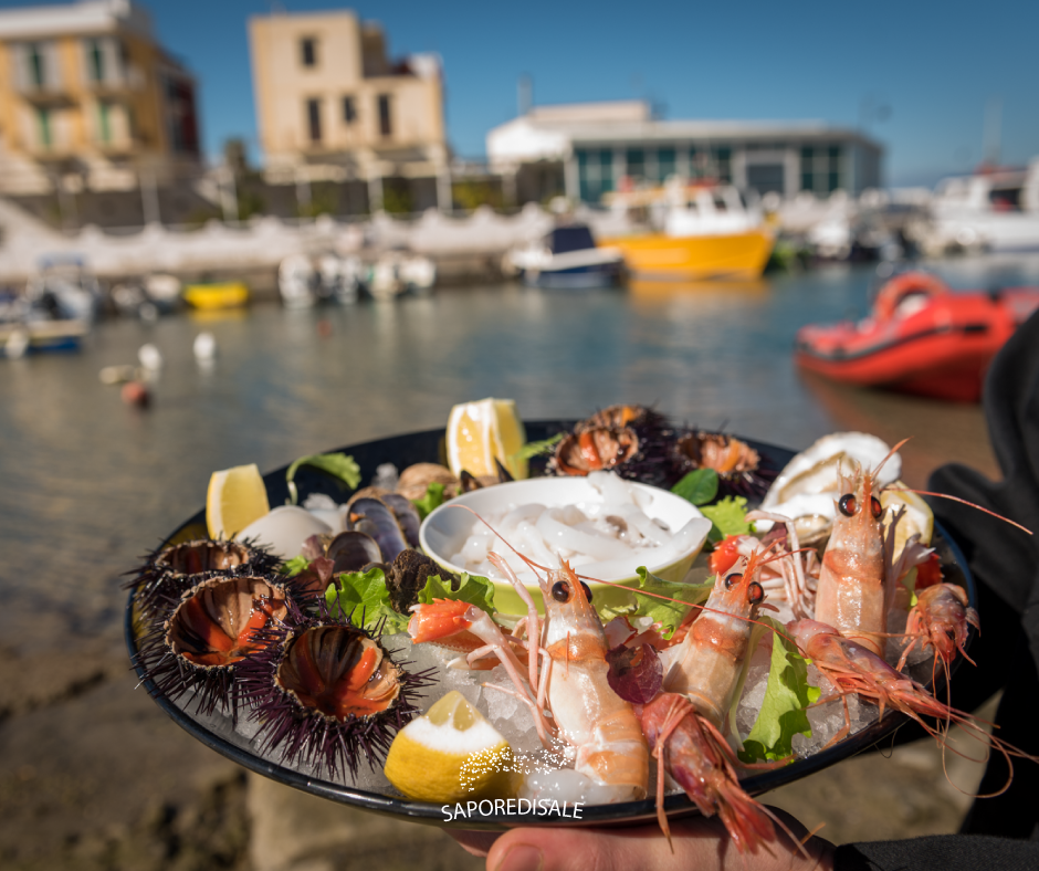Piatto di frutti di mare con gamberi, ricci di mare e fette di limone, servito vicino a un porto con barche ed edifici.