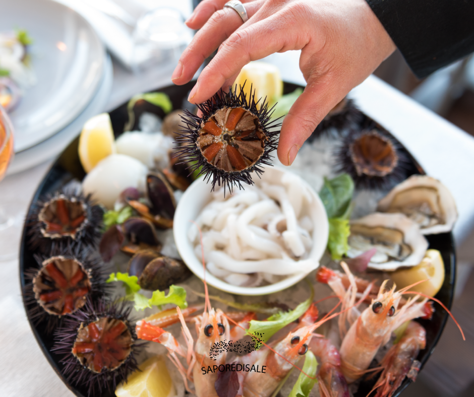 Mano che tiene un riccio di mare sopra un piatto di frutti di mare, tra cui gamberi, ostriche e calamari.