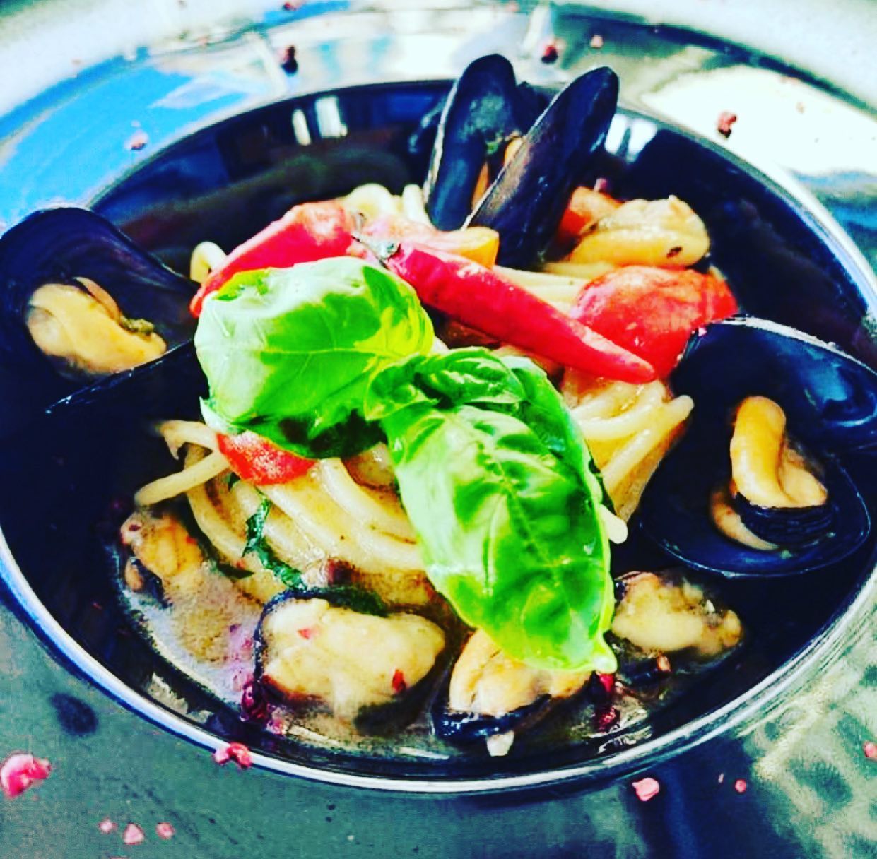 Pasta con cozze, pomodori, peperoncino e basilico, servita in una ciotola scura su un piatto d'argento.
