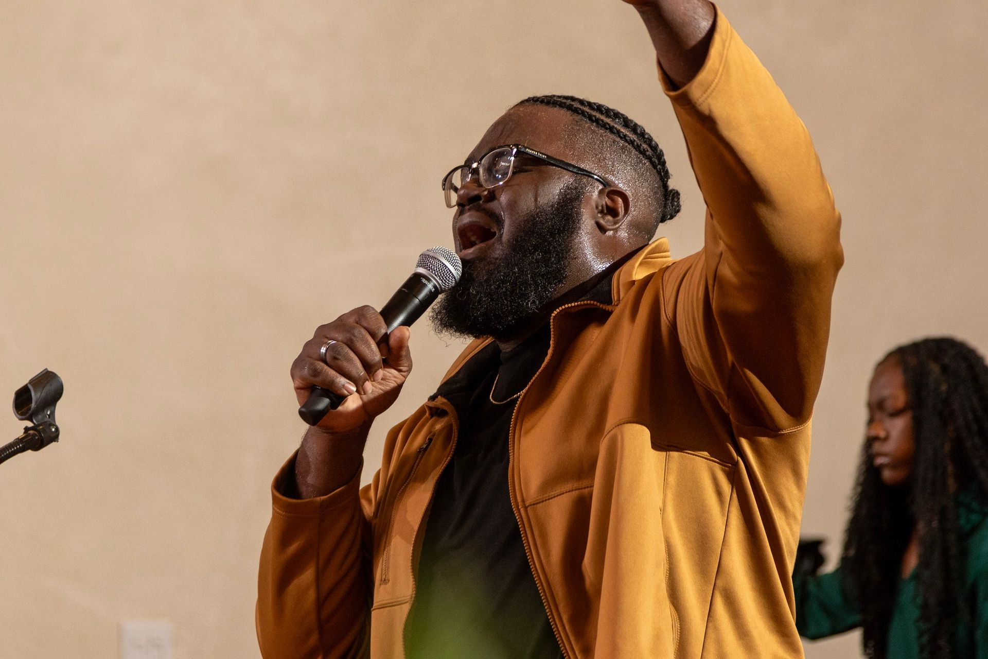 Man speaking into a microphone with one arm raised, wearing a mustard jacket on a stage