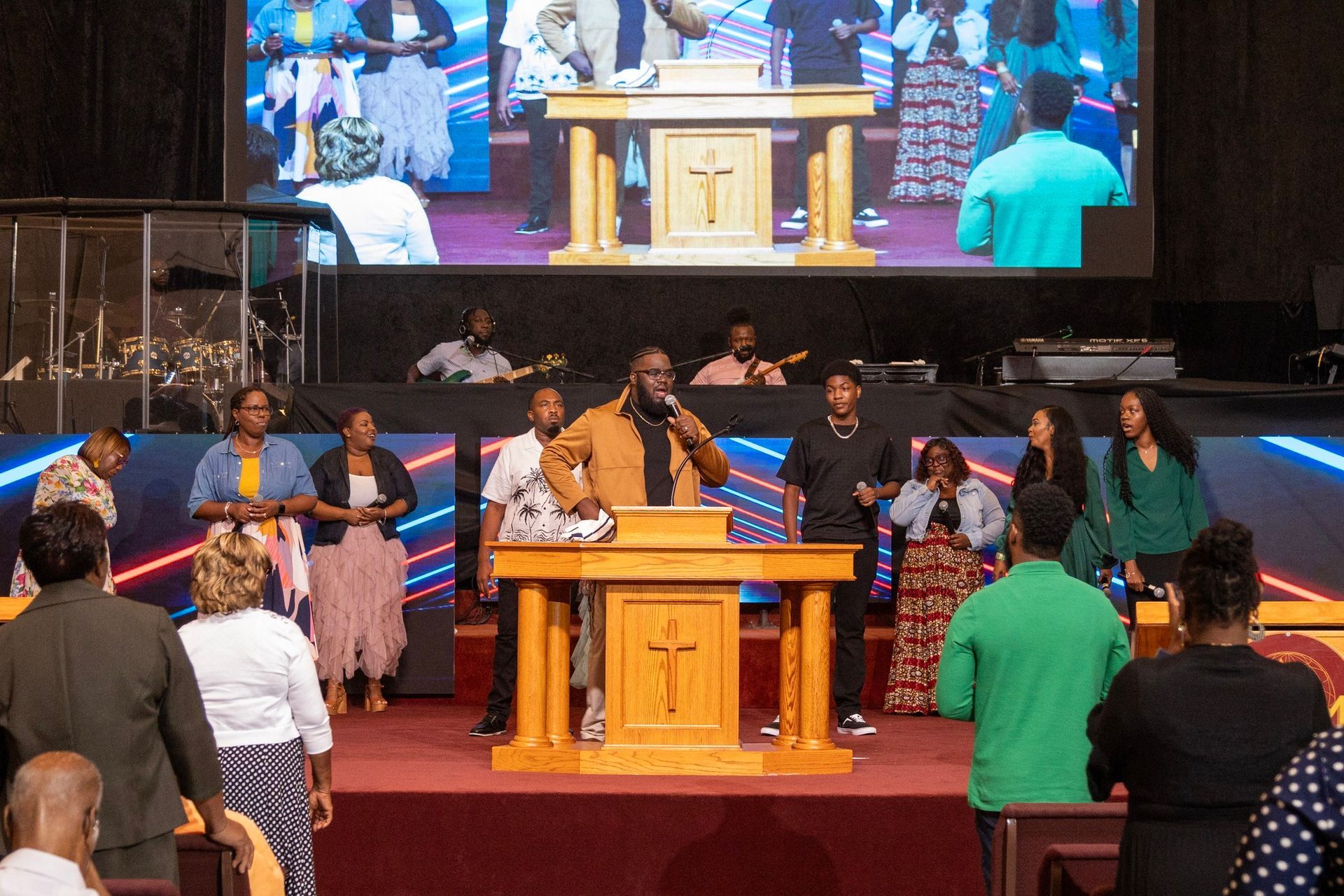 Church service with people on stage singing behind a wooden podium, projected on a large screen.