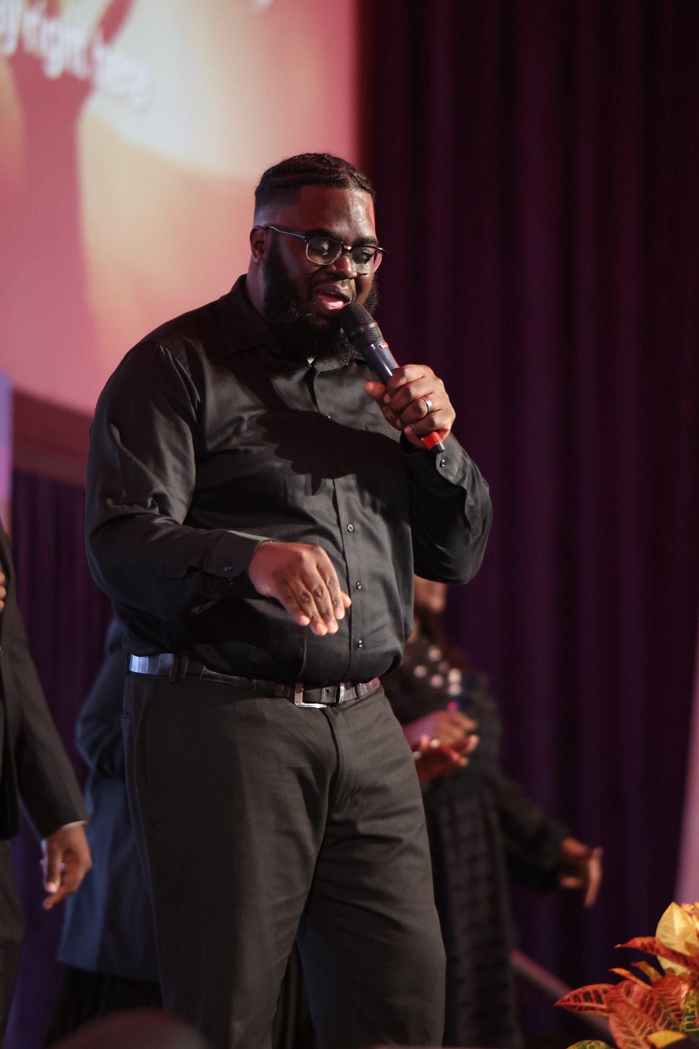 Man in black shirt speaking into a microphone on a stage with purple curtains and warm lighting