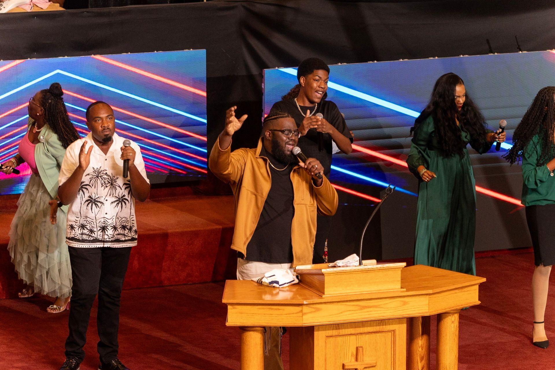 Worship team singing on stage with microphones and raised hands, led by a speaker at a wooden podium