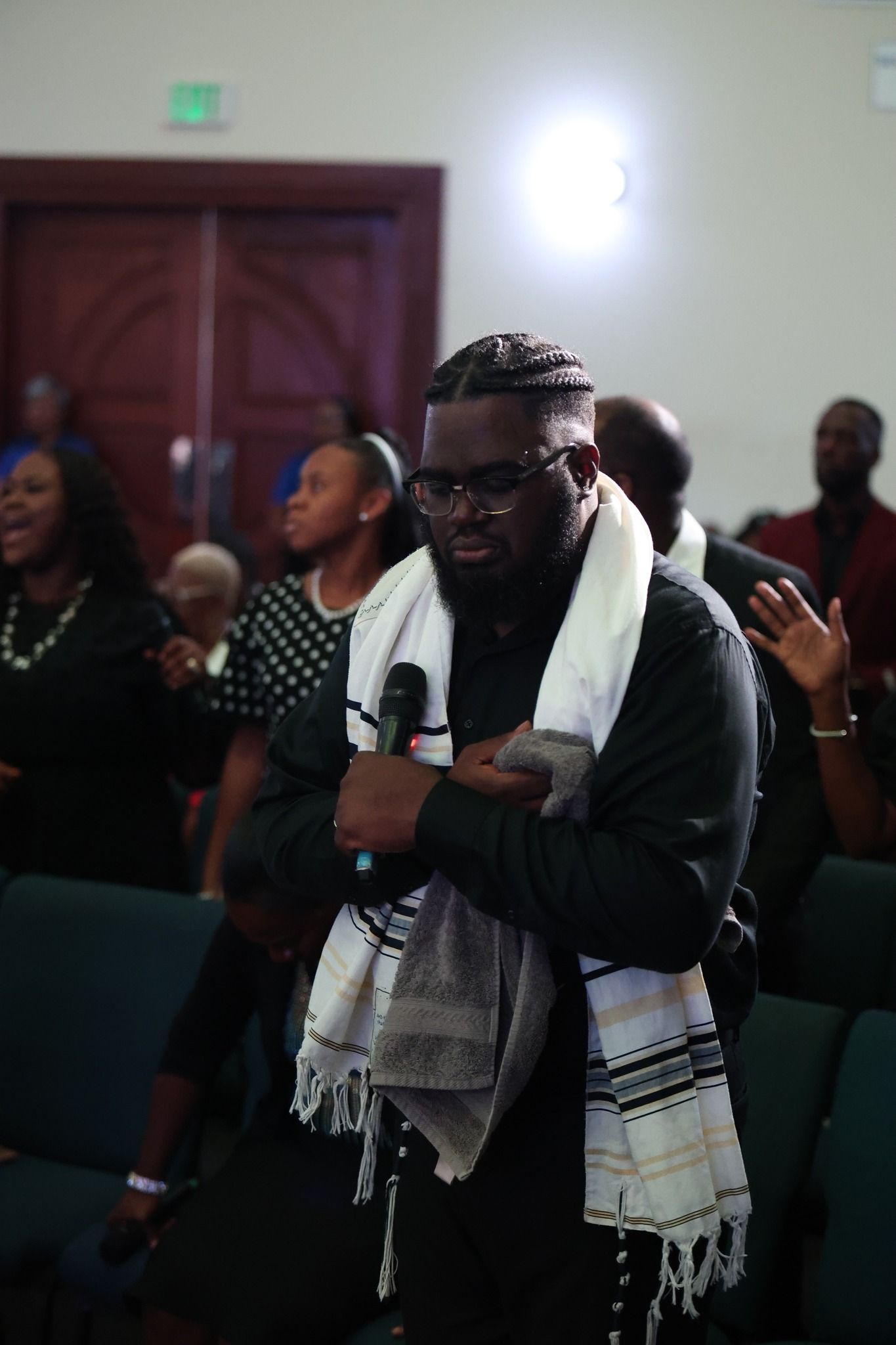 Man standing in a church aisle with arms crossed and a white scarf, among seated attendees